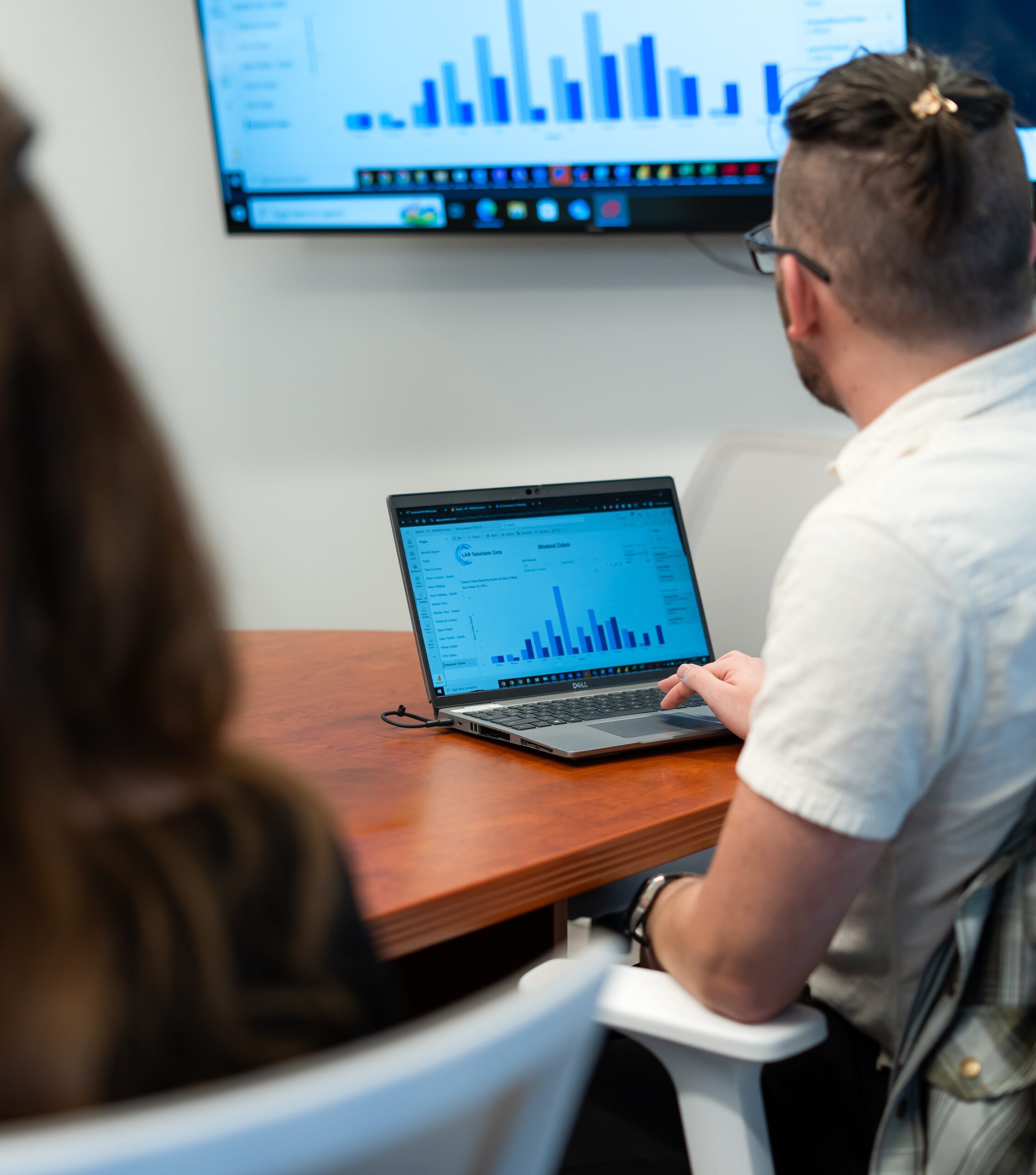 A person reviewing bar chart data on a laptop during a meeting, with the same chart displayed on a large screen.